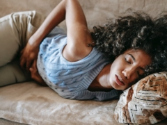 High angle of African American woman with afro hairstyle and closed eyes having pain in belly lying on sofa