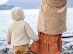 Anonymous baby holding skirt of crop woman near river in winter countryside