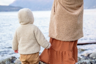 Anonymous baby holding skirt of crop woman near river in winter countryside